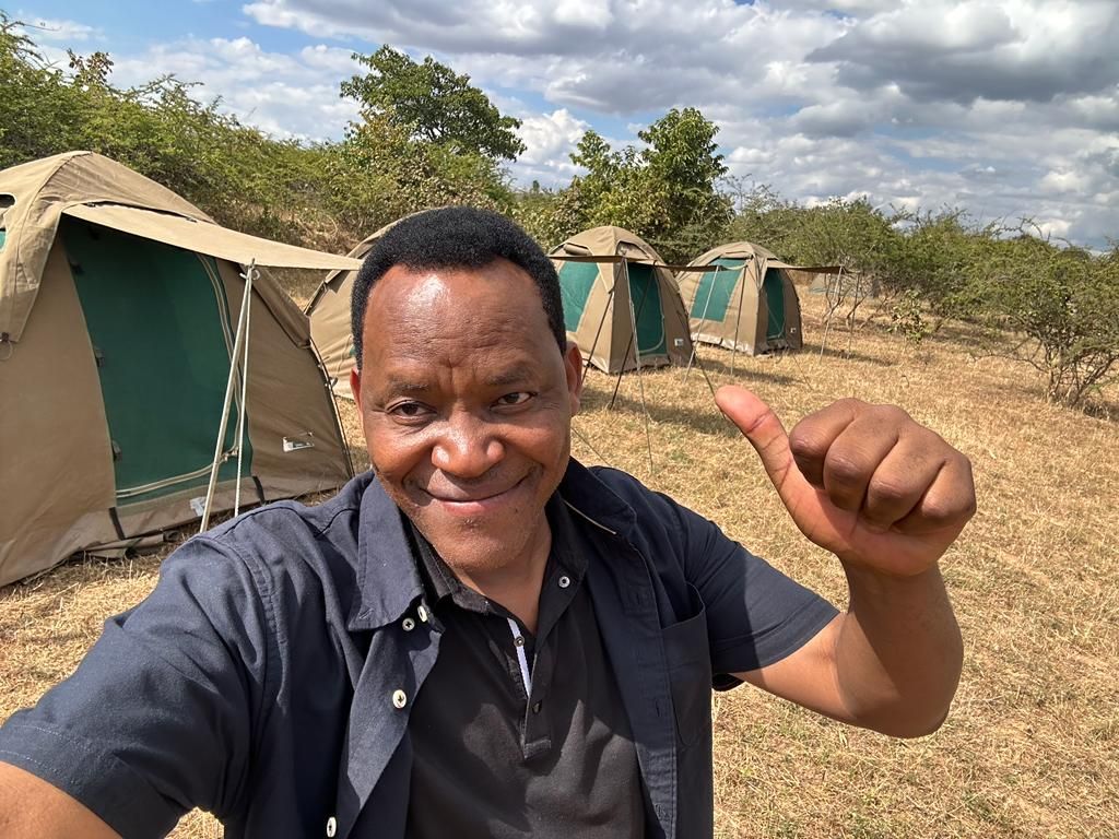A smiling man giving a thumbs-up in front of a row of clinic tents in the bush