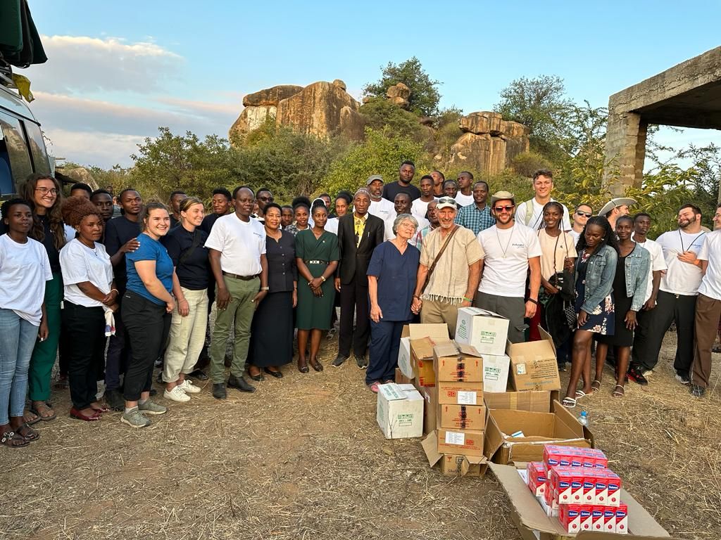 A large group of Jitolee volunteers and community members standing together beside boxes of donated supplies, with rocky hills behind them